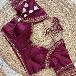 May include: A maroon blouse with intricate gold embroidery along the neckline, sleeves, and hem. The blouse features a V-neck and short sleeves. The garment is displayed on a woven, circular mat, with a sprig of white flowers.