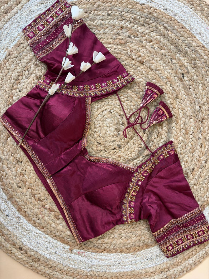 May include: A maroon blouse with intricate gold embroidery along the neckline, sleeves, and hem. The blouse features a V-neck and short sleeves. The garment is displayed on a woven, circular mat, with a sprig of white flowers.