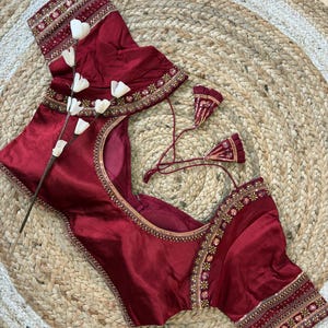 May include: A maroon-colored blouse with intricate embroidery along the neckline and sleeves. The blouse features a scoop neck and short sleeves. The embroidery includes gold and other colorful accents. The blouse is displayed on a woven, circular rug.