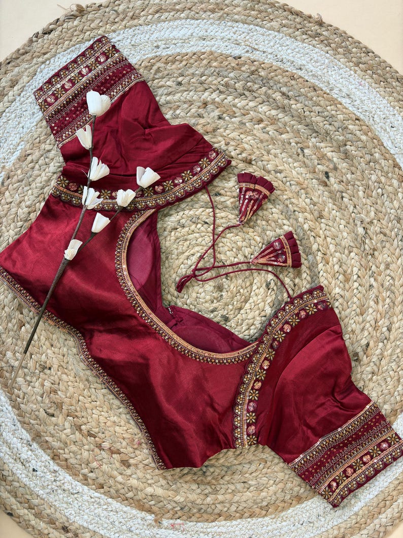 May include: A maroon-colored blouse with intricate embroidery along the neckline and sleeves. The blouse features a scoop neck and short sleeves. The embroidery includes gold and other colorful accents. The blouse is displayed on a woven, circular rug.
