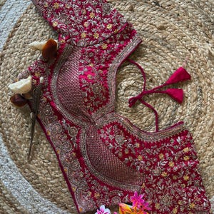 以下が含まれることがあります： マゼンタ色の、花柄と装飾が施された刺繍が施されたブラウス。ハートネックラインと半袖で、ビーズ細工と装飾的なトリムが施されています。フォーマルウェアのデザインです。