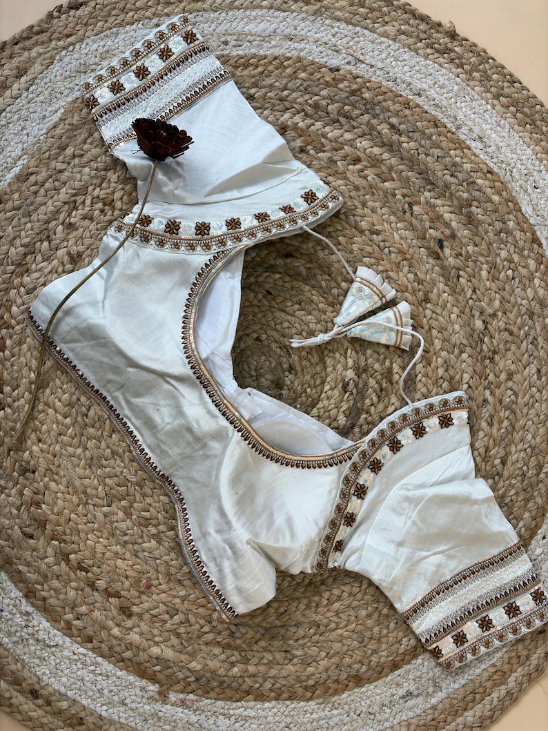 May include: Ivory-colored blouse with intricate brown and gold embroidery along the neckline, sleeves, and edges. The blouse features a scoop neckline and short sleeves. The garment is displayed on a woven, circular mat.