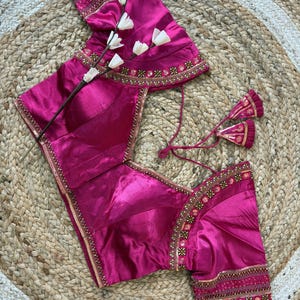 May include: A magenta-colored blouse with intricate gold embroidery along the neckline, sleeves, and hem. The blouse features a V-neck and short sleeves. The garment is displayed on a woven, neutral-toned surface, with a sprig of white flowers.