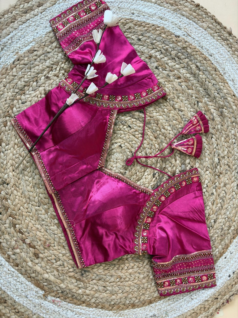 May include: A magenta-colored blouse with intricate gold embroidery along the neckline, sleeves, and hem. The blouse features a V-neck and short sleeves. The garment is displayed on a woven, neutral-toned surface, with a sprig of white flowers.