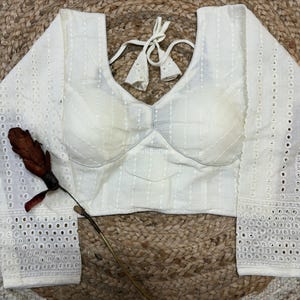 Puede incluir: Blusa blanca de manga larga con detalles de ojal en las mangas y escote en V. La blusa tiene un cierre de lazo en la espalda y está hecha de una tela texturizada. Una flor seca con un acento rojo se coloca cerca de la blusa.