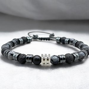 May include: A beaded bracelet featuring a combination of dark gray and black beads, including lava rock and hematite. The bracelet has a silver-colored central accent and an adjustable cord closure. This accessory is suitable for everyday wear.