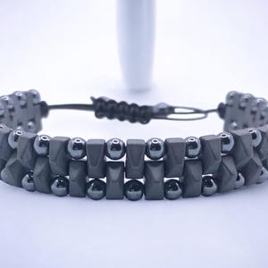 May include: A dark gray bracelet with a double-strand design. The bracelet features geometric-shaped beads and small, round beads. It has an adjustable black cord closure.