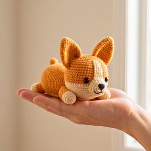 Puede incluir: Un juguete de perro corgi tejido a ganchillo en tonos naranja, crema y blanco. El peluche hecho a mano tiene orejas grandes y erguidas, una pequeña cola y ojos negros de botón. El corgi está tumbado, con las patas extendidas.