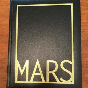 May include: A black hardcover book with a gold rectangular border on the front cover. The word MARS is printed in large gold capital letters at the bottom of the cover, integrated into the border design. A small orange ribbon bookmark is visible.