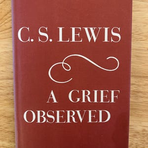 May include: A book titled "A Grief Observed" by C.S. Lewis. The cover is a deep red with white text and a decorative swirl. The book is rectangular and appears to be in good condition.