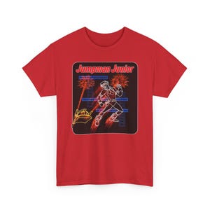 Puede incluir: Camiseta roja con un gráfico que dice "Jumpman Junior". El gráfico muestra un contorno de neón de una figura corriendo, con un paisaje urbano y fuegos artificiales. El diseño está dentro de un marco rectangular.
