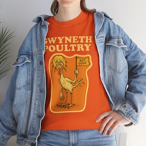 Puede incluir: Camiseta naranja con el texto "SWYNETH POULTRY" y un dibujo de un pollo sosteniendo una bandera que dice "BUY GOOF". La camiseta se lleva con una chaqueta vaquera azul.