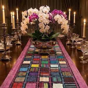 May include: A formal dining table is set with a centerpiece of white and purple orchids in a gold-toned bowl. Lit candles in ornate holders flank the floral arrangement. A colorful patchwork runner adds a vibrant contrast to the dark wood table.