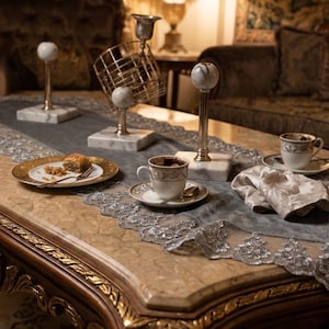 May include: A luxurious coffee table setting with ornate gold trim. The table is adorned with decorative marble and gold accents, including candle holders and serving dishes. A gray runner with silver lace trim complements the setting, with coffee cups and a pastry on a gold plate.