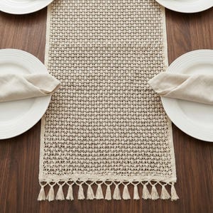 Pode incluir: Caminho de mesa creme com um design geométrico vazado e uma franja de borlas. O caminho de mesa está colocado sobre uma mesa de madeira escura, com pratos brancos e guardanapos dobrados de cada lado.