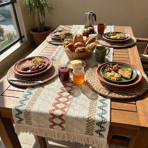 Op de afbeelding: Een houten tafel gedekt voor het ontbijt met borden met eten, brood en drankjes. De tafel is bedekt met een patroonloper en placemats. Potten jam en honing, samen met mokken en een koffiepot, maken de scène compleet.