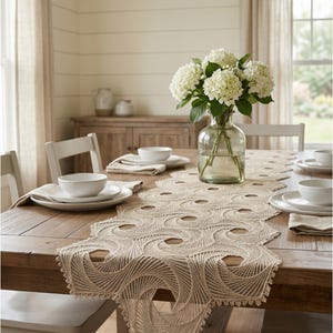 May include: A dining table set for a meal, featuring a beige, intricately patterned table runner. White plates, bowls, and napkins are set for each place setting. A clear glass vase holds a bouquet of white hydrangeas, adding a touch of freshness.