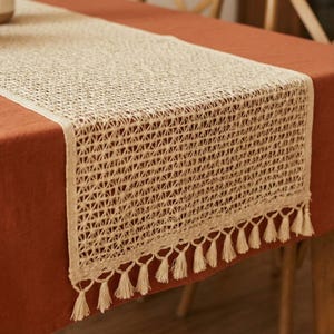 Op de afbeelding: Een beige geweven tafelloper met een geometrisch patroon en een franje van kwastjes langs de onderrand. De loper ligt op een roestkleurig tafelkleed, wat een contrast in texturen en kleuren creëert. Dit decoratieve stuk voegt elegantie toe.