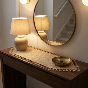 May include: A warm-toned entryway scene featuring a dark wood console table. Two beige lamps with linen shades sit on the table, alongside a patterned runner with tassels. A gold-toned bowl holds keys, and a round mirror hangs above.