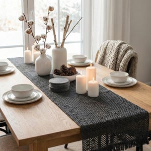 May include: A dining table set for a meal. A dark gray table runner with fringe is centered on the wooden table. White plates, bowls, and candles are arranged on the table. Vases with cotton stems and birch branches are also present.