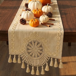 May include: A beige table runner with a decorative crochet design and tassels, adorned with white and orange pumpkins, pine cones, and cinnamon sticks. The runner is placed on a dark wooden table, creating a fall-themed display.