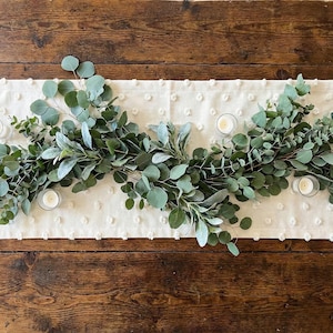 May include: A table runner with a cream-colored, dotted pattern, adorned with a garland of green eucalyptus and sage leaves. Small glass votive candles are evenly spaced along the runner, creating a natural, elegant centerpiece.
