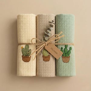 May include: A set of three rolled waffle-weave towels in cream, tan, and sage green, tied with twine and a sprig of greenery. Each towel is embroidered with a cactus in a terracotta pot. A small tag reads "Elia Goods".
