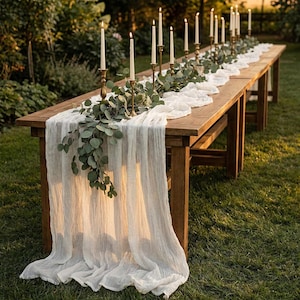 Puede incluir: Una larga mesa de madera preparada para un evento al aire libre. La mesa está adornada con un camino de mesa blanco, vegetación, candelabros dorados y altas velas blancas. La escena está ambientada en un césped, con la luz del sol iluminando el arreglo.