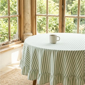 May include: A round table with a green and white striped tablecloth and a ruffled edge. A white mug with a dark drink sits on the table. The table is in front of a window with a view of greenery.