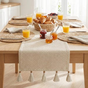 Könnte beinhalten: Ein Holztisch, der für das Frühstück gedeckt ist. Auf dem Tisch befinden sich gewebte Tischsets, Orangensaftgläser, ein Korb mit Gebäck und kleine Gläser mit Marmelade und Honig. Ein cremefarbener Tischläufer mit Quasten verläuft in der Mitte.