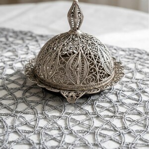May include: A silver filigree butter dish with a domed lid and ornate handle. The dish sits on a matching scalloped base, showcasing intricate openwork patterns. The decorative piece is a beautiful addition to any table setting.