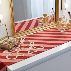 May include: A vanity table with a red and pink striped runner, a gold-framed mirror, and various perfume bottles. A glass jewelry box and decorative white floral embroidery are on the table. The scene is well-lit and features a clean, organized aesthetic.