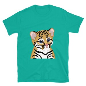 May include: A teal t-shirt with a pixelated illustration of a baby ocelot. The ocelot has tan fur with black stripes, blue eyes, and a pink nose. The shirt is made of a soft material and has short sleeves.