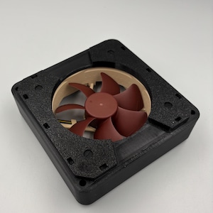 May include: A computer fan with a black frame and a brown fan blade. The fan has a square shape and is designed to cool computer components. The fan blades are a reddish-brown color, and the frame has a textured surface.
