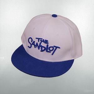 Può includere: Un cappellino da baseball bianco e blu con la scritta "THE SANDLOT" ricamata in blu. Il cappellino ha una visiera piatta e un bottone sulla parte superiore. Un cappello in stile classico.