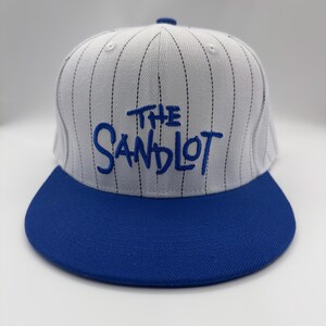 Può includere: Cappellino da baseball con corona a righe bianche e grigie e visiera blu. La scritta "THE SANDLOT" è ricamata in blu sul davanti. Il cappellino ha una visiera piatta e un bottone sulla parte superiore.