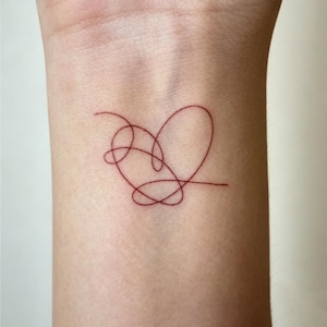 May include: A small, red heart tattoo on a wrist. The design is a continuous line forming a heart shape with intertwined loops. The tattoo is a minimalist design, with a simple, elegant aesthetic.