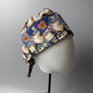 Può includere: Un cappello chirurgico blu fantasia con un motivo ripetuto di gatti dei cartoni animati, fiocchi e stelle. Le parole "Happy" e "Cute" sono stampate anche sul cappello. Il cappello è legato sul retro ed è esposto su una testa di manichino.