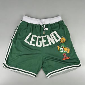 Könnte beinhalten: Grüne Mesh-Shorts mit weißem Besatz und weißem Bund. Das Wort "LEGEND" steht in großen weißen Buchstaben auf der Vorderseite. Ein Cartoon-Vogel mit einem grünen Trikot mit der Nummer 3 befindet sich auf dem rechten Bein.