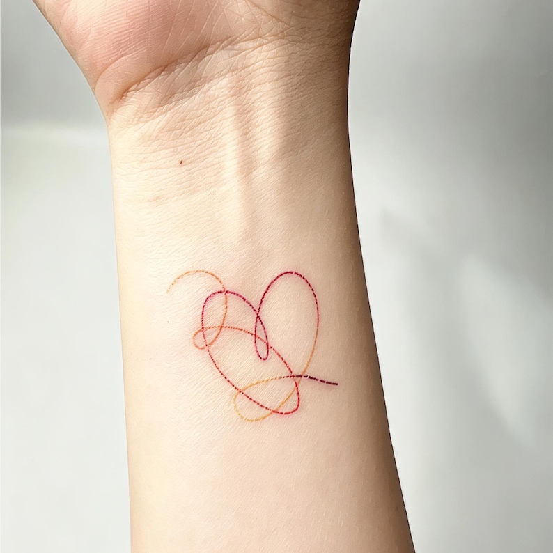 May include: A red and gold heart-shaped tattoo on a wrist. The design features a continuous line forming a heart shape, with additional looping lines. The tattoo is a minimalist design, with a delicate and intricate appearance.