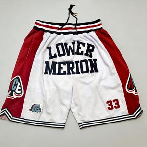 Può includere: Pantaloncini da basket in rete bianca con pannelli laterali rossi. I pantaloncini presentano la scritta "LOWER MERION" in blu navy, il numero "33" in rosso e una grafica bulldog. Rifinitura a righe bianche e nere.