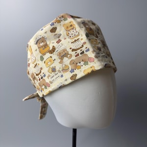Può includere: Un cappello chirurgico giallo chiaro con un motivo ripetuto di orsetti dei cartoni animati in abbigliamento da chef, prodotti da forno e la scritta "Baker Bear Love Cookies". Il cappello è legato sul retro e mostrato su una testa di manichino.