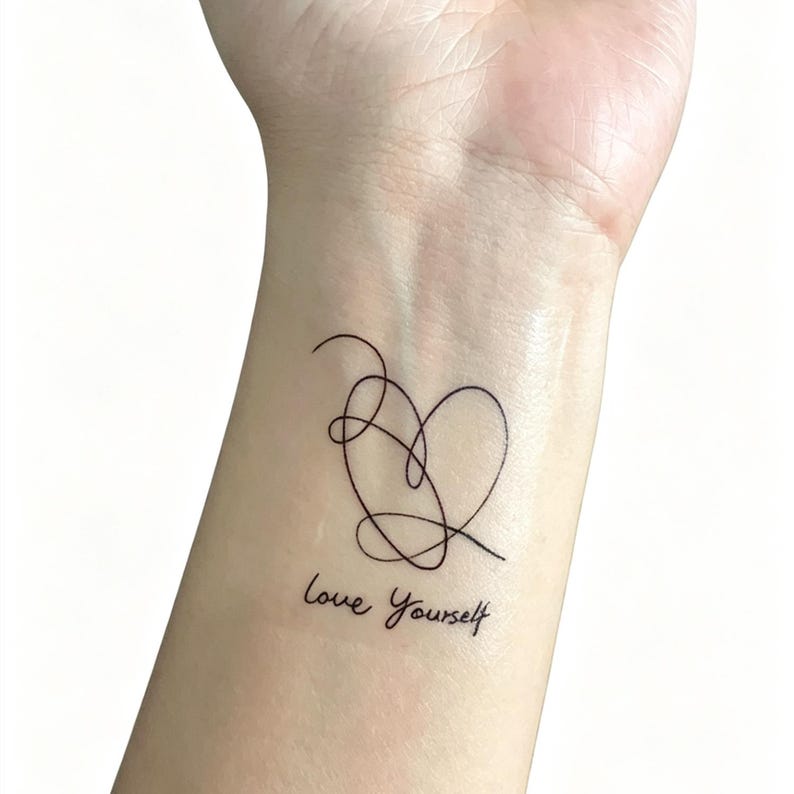 May include: A black line art tattoo of a heart shape with the words "Love Yourself" in cursive script. The tattoo is on a person's wrist.