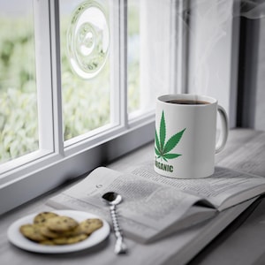 Könnte beinhalten: Eine weiße Keramiktasse mit einem grünen Cannabisblatt-Design und dem Wort "ORGANIC" in Grün. Die Tasse steht auf einem offenen Buch neben einem Teller mit Keksen und einem Löffel. Dampf steigt aus der Tasse auf.