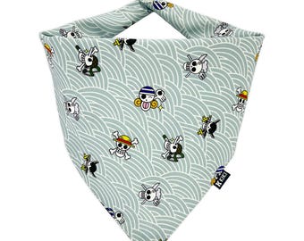 May include: A light blue dog bandana with a repeating pattern of cartoon skulls and crossbones, and other character graphics. The bandana has a wave-like design in white. A small black tag with white text is attached to the bandana.