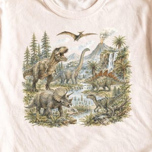 以下が含まれることがあります： 淡いピーチカラーのTシャツで、先史時代の風景の中に様々な恐竜が描かれた詳細なグラフィックプリントが施されています。シーンには、ティラノサウルス、トリケラトプス、首の長い恐竜、ステゴサウルス、そして火山の近くを飛ぶプテロダクティルスが含まれています。