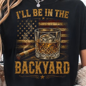May include: Black t-shirt with a graphic design featuring an American flag, a glass of whiskey with ice, and a cigar. The text on the shirt reads "I'll be in the backyard."