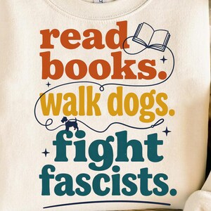 Könnte beinhalten: Ein cremefarbenes Sweatshirt mit Rundhalsausschnitt und einem Grafikdruck mit dem Text "read books. walk dogs. fight fascists.". Das Design enthält ein kleines Symbol eines offenen Buches, eine Hundesilhouette an der Leine und dekorative Sterne.
