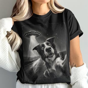 Puede incluir: Una camiseta negra de manga corta con un estampado gráfico de un perro tomándose un selfie. La ilustración en blanco y negro muestra a un perro en primer plano con un OVNI sobrevolando el cielo nocturno sobre un camino rural con luz.