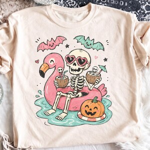 May include: A cream-colored t-shirt featuring a graphic of a skeleton wearing heart-shaped sunglasses, sitting in a pink flamingo pool float in turquoise water. The skeleton holds two coconut drinks, while a jack-o'-lantern and two bats appear.